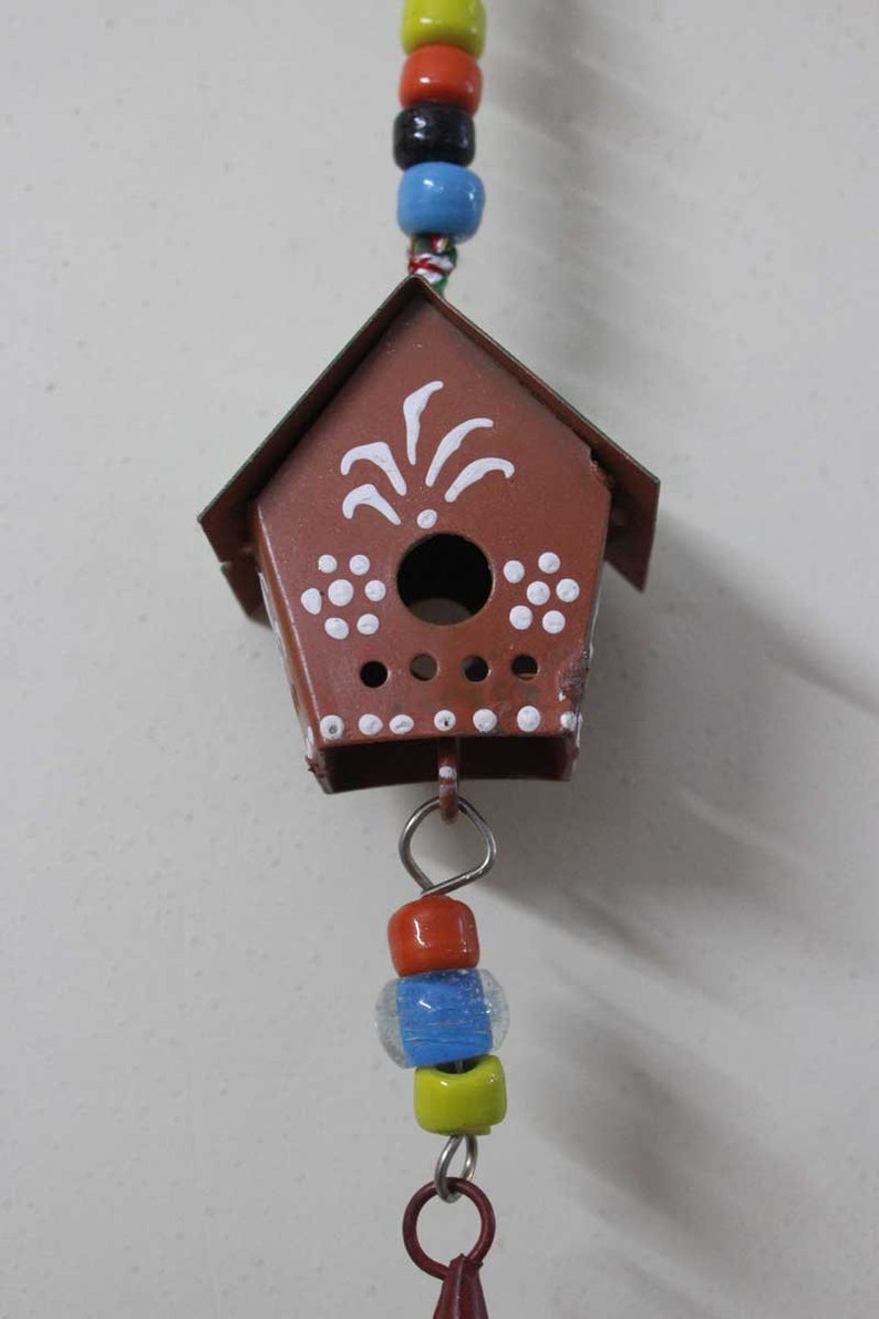Puede incluir: Una casita de p&aacute;jaros de metal marr&oacute;n con detalles pintados en blanco cuelga de una cuerda de cuentas de colores. La casita de p&aacute;jaros tiene una peque&ntilde;a abertura redonda y un dise&ntilde;o decorativo en la parte delantera.