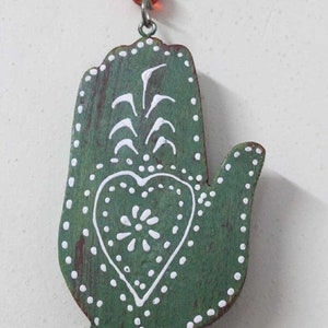 Puede incluir: Mano de madera verde con dise&ntilde;os pintados en blanco, incluyendo un coraz&oacute;n y una flor estilizada. La mano est&aacute; unida a una cuerda de cuentas de vidrio azules y rojas.
