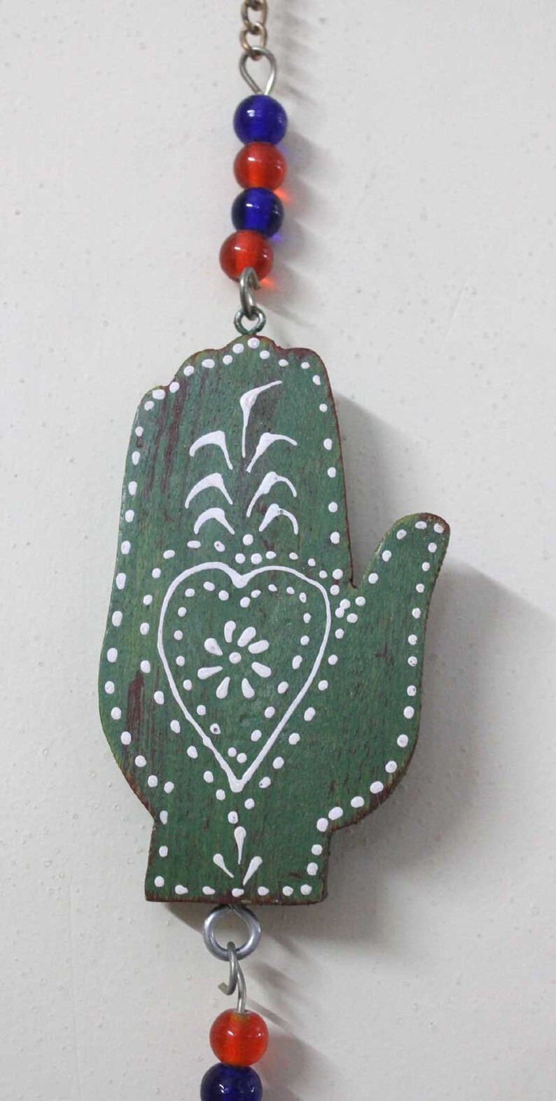Puede incluir: Mano de madera verde con dise&ntilde;os pintados en blanco, incluyendo un coraz&oacute;n y una flor estilizada. La mano est&aacute; unida a una cuerda de cuentas de vidrio azules y rojas.