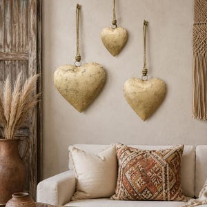 May include: Three heart-shaped wall decorations with a distressed gold finish, each suspended by a thick, natural-colored rope loop. The hearts vary in size and are arranged vertically and horizontally on the wall above a cream-colored sofa.