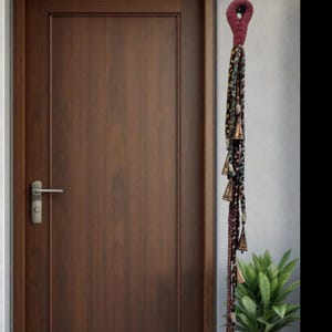 Puede incluir: Un adorno decorativo para puerta con un lazo burdeos y una larga hebra de cuerda trenzada. La cuerda está adornada con pequeñas campanas de latón y cuentas de colores. La puerta es de color marrón oscuro.