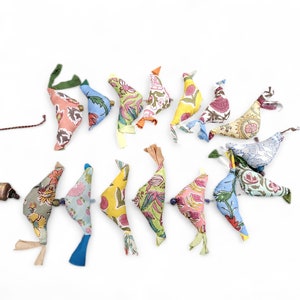 May include: A collection of colorful fabric bird ornaments with floral patterns, arranged on a white background. Each bird has a unique color scheme, including pink, blue, yellow, and green, with ribbon and bead accents. A small bottle and string are also present.