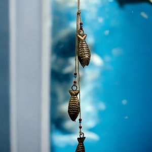 May include: A string of three gold fish-shaped ornaments hanging from a brown string with colourful beads. The ornaments are hanging against a blue background.
