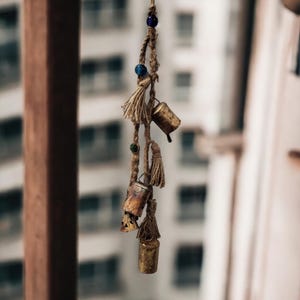 以下が含まれることがあります： 青いビーズと緑のビーズが付いた茶色のロープから、小さな金のベルと茶色のタッセルがぶら下がっています。ベルは、建物のぼやけた背景の前にぶら下がっています。