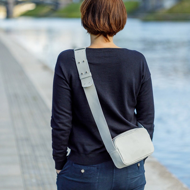 K&ouml;nnte beinhalten: Eine Person tr&auml;gt ein schwarzes Langarmshirt und eine blaue Jeans mit einer hellgrauen Umh&auml;ngetasche mit langem Riemen.