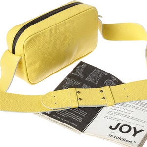 K&ouml;nnte beinhalten: Eine gelbe Leder-Umh&auml;ngetasche mit schwarzem Rei&szlig;verschluss und einem langen verstellbaren Riemen. Die Tasche liegt auf einem Buch mit dem Text "JOY and revolution."