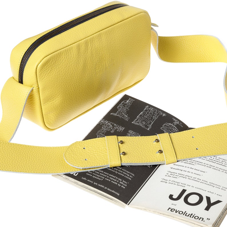 K&ouml;nnte beinhalten: Eine gelbe Leder-Umh&auml;ngetasche mit schwarzem Rei&szlig;verschluss und einem langen verstellbaren Riemen. Die Tasche liegt auf einem Buch mit dem Text "JOY and revolution."
