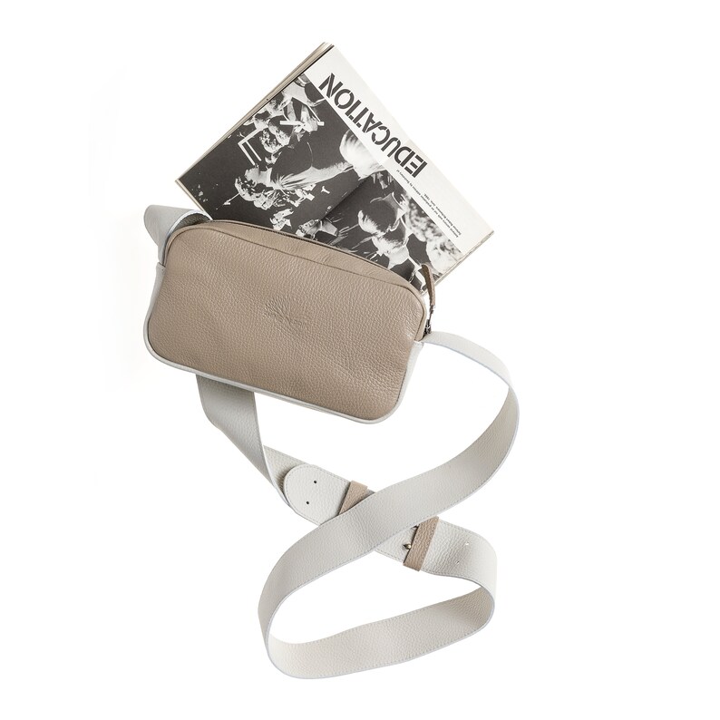 K&ouml;nnte beinhalten: Eine beige Leder-Umh&auml;ngetasche mit einem wei&szlig;en, verstellbaren Trageriemen. Die Tasche ist offen und ein Magazin mit dem Titel "EDUCATION" ist teilweise im Inneren sichtbar.