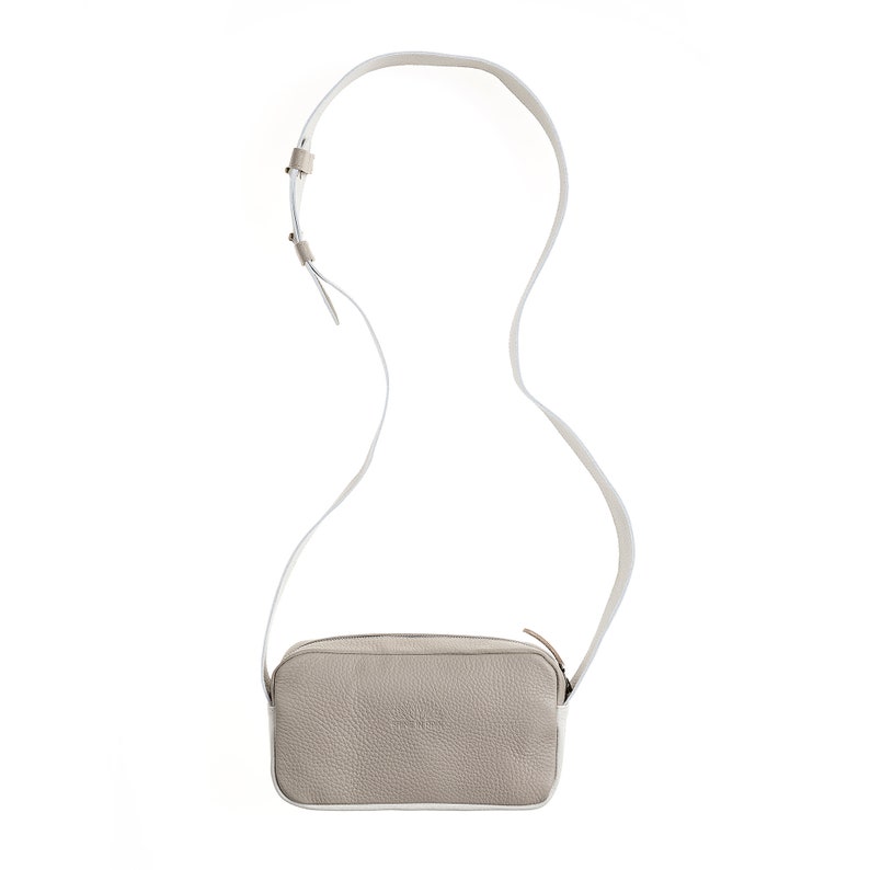 K&ouml;nnte beinhalten: Eine beige Leder-Umh&auml;ngetasche mit einem wei&szlig;en Trageriemen. Die Tasche hat eine rechteckige Form und einen Rei&szlig;verschluss.