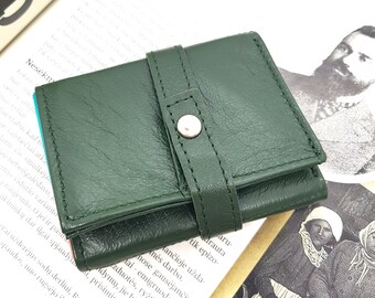 May include: A dark green leather wallet with a fold-over flap and a silver button closure. The wallet is closed and lying on a book.