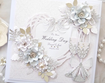 May include: A white wedding card with a heart-shaped design, silver and gold embellishments, and the text "On Your Wedding Day David & Jade 10th June 2024".