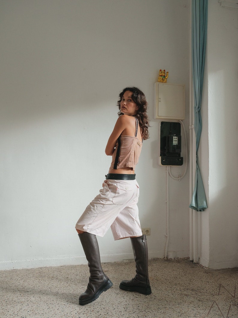 May include: A woman wearing a brown tank top with a black lace trim, white shorts, and brown knee-high boots. She is standing in a room with white walls and a blue curtain.