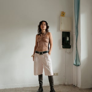 May include: A woman wearing a brown halter top with a silver belt, white shorts, and black knee-high boots. She is standing in a room with white walls and a light blue curtain.
