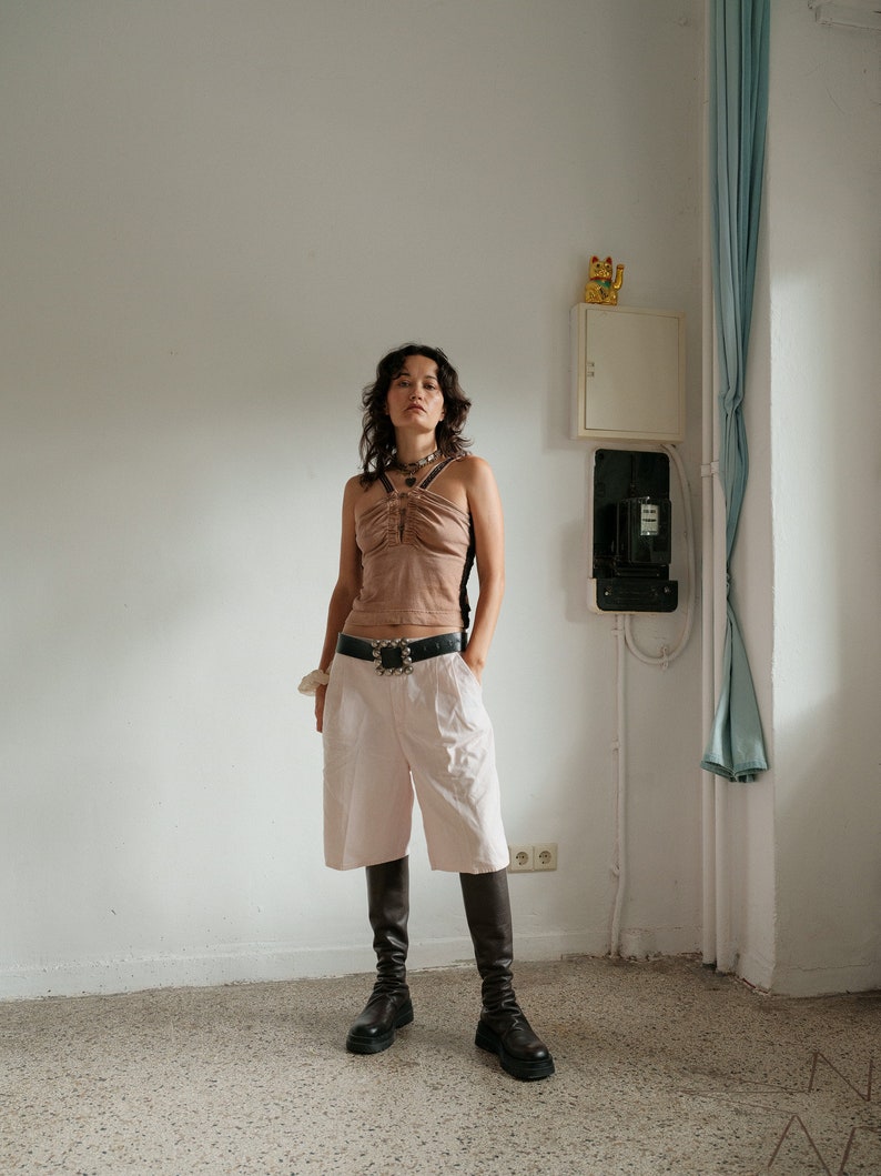 May include: A woman wearing a brown halter top with a silver belt, white shorts, and black knee-high boots. She is standing in a room with white walls and a light blue curtain.