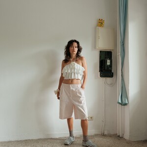 May include: A woman wearing a white ruffled halter top, pink cargo shorts, and white platform sandals. She is standing in a room with white walls and a light blue curtain.