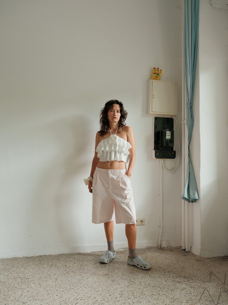 May include: A woman wearing a white ruffled halter top, pink cargo shorts, and white platform sandals. She is standing in a room with white walls and a light blue curtain.