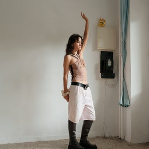 May include: A woman wearing a light brown tank top with a black trim, white pants, and black boots. She is standing in a room with a white wall and a white electrical box.