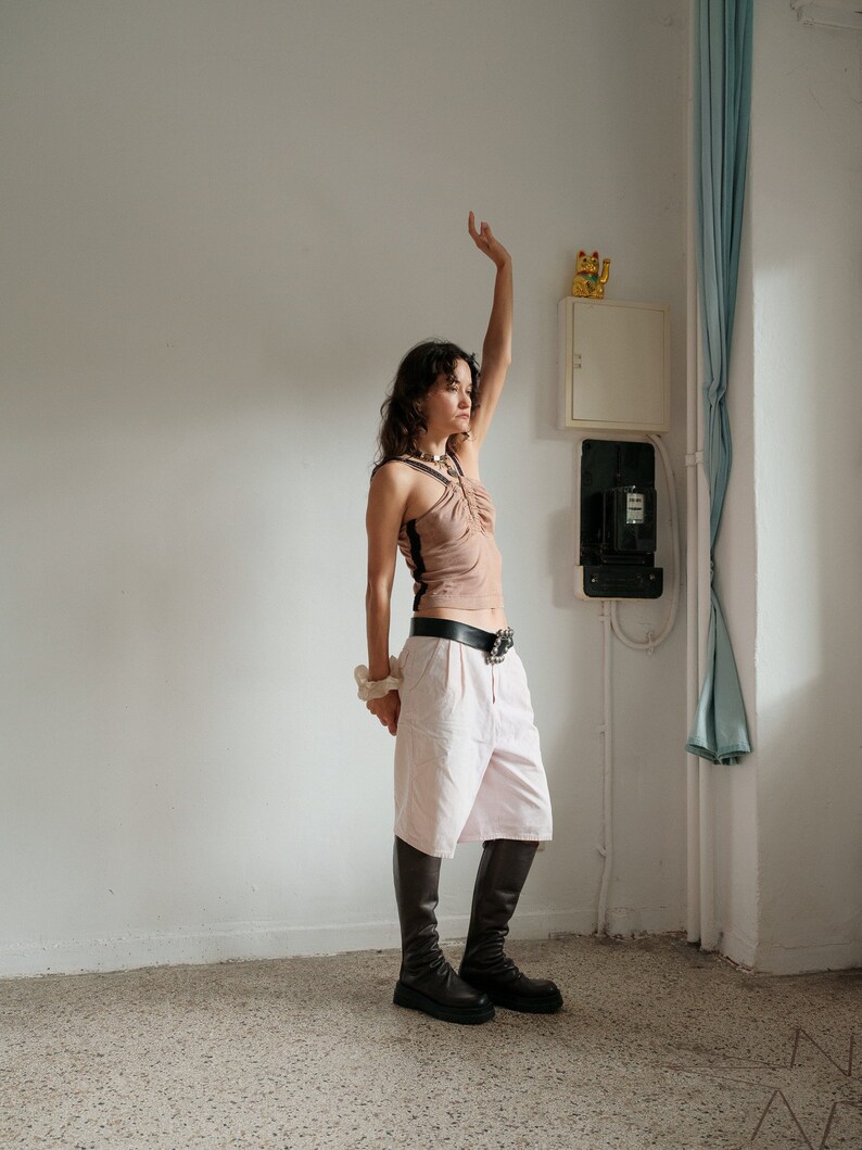 May include: A woman wearing a light brown tank top with a black trim, white pants, and black boots. She is standing in a room with a white wall and a white electrical box.