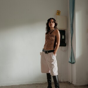May include: A woman wearing a brown halter top, a pink pair of shorts, and black knee-high boots. She is standing in a room with a white wall and a blue curtain.