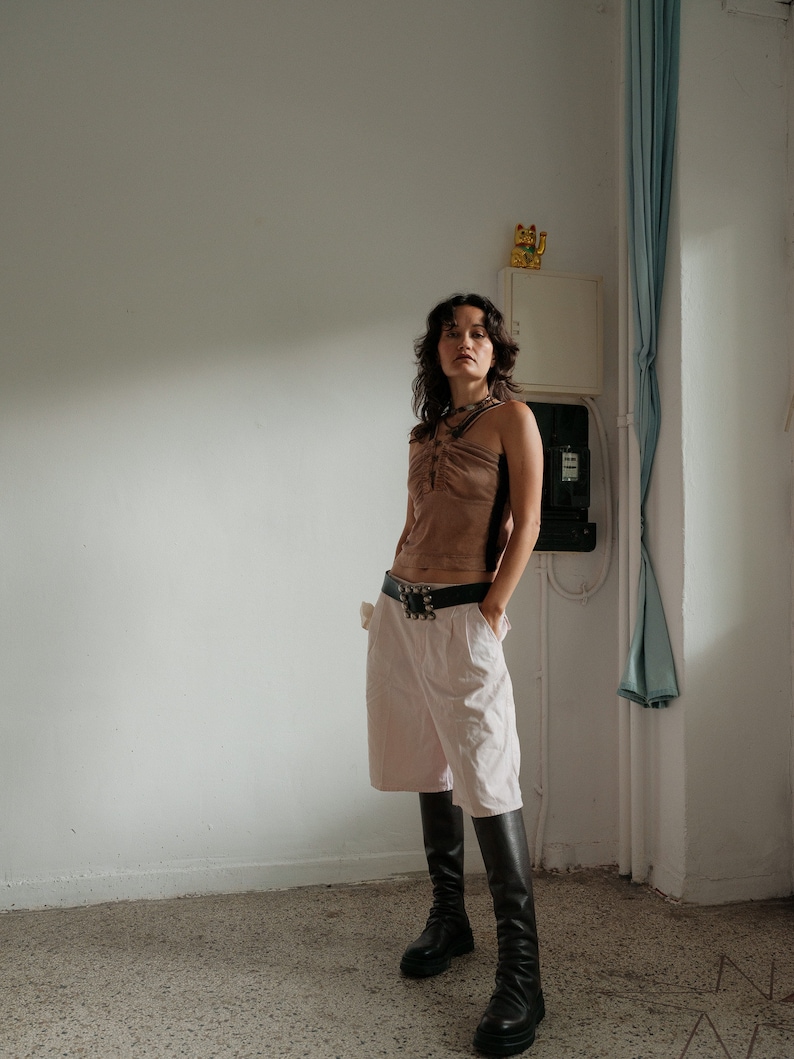 May include: A woman wearing a brown halter top, a pink pair of shorts, and black knee-high boots. She is standing in a room with a white wall and a blue curtain.