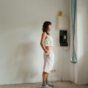 May include: A woman wearing a white ruffled top and pink shorts stands in a room with white walls and a blue curtain. She is wearing gray clogs and looking to the right.
