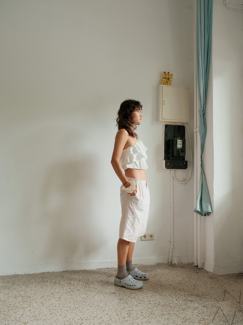 May include: A woman wearing a white ruffled top and pink shorts stands in a room with white walls and a blue curtain. She is wearing gray clogs and looking to the right.