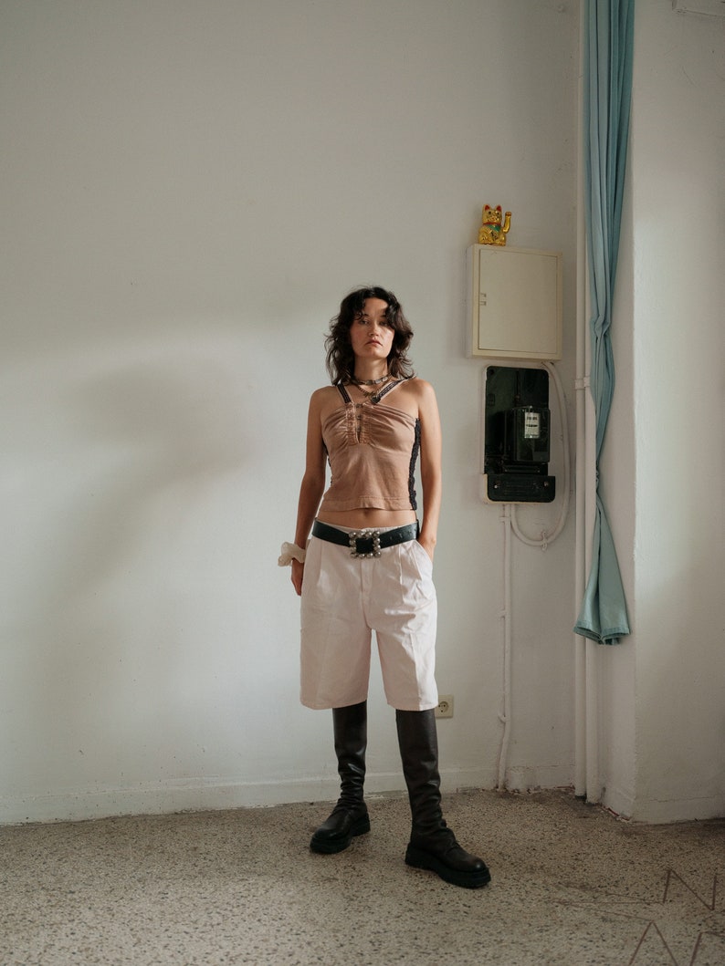 May include: A woman wearing a brown halter top with a black belt, white shorts, and black knee-high boots. She is standing in a room with a white wall and a white cabinet.