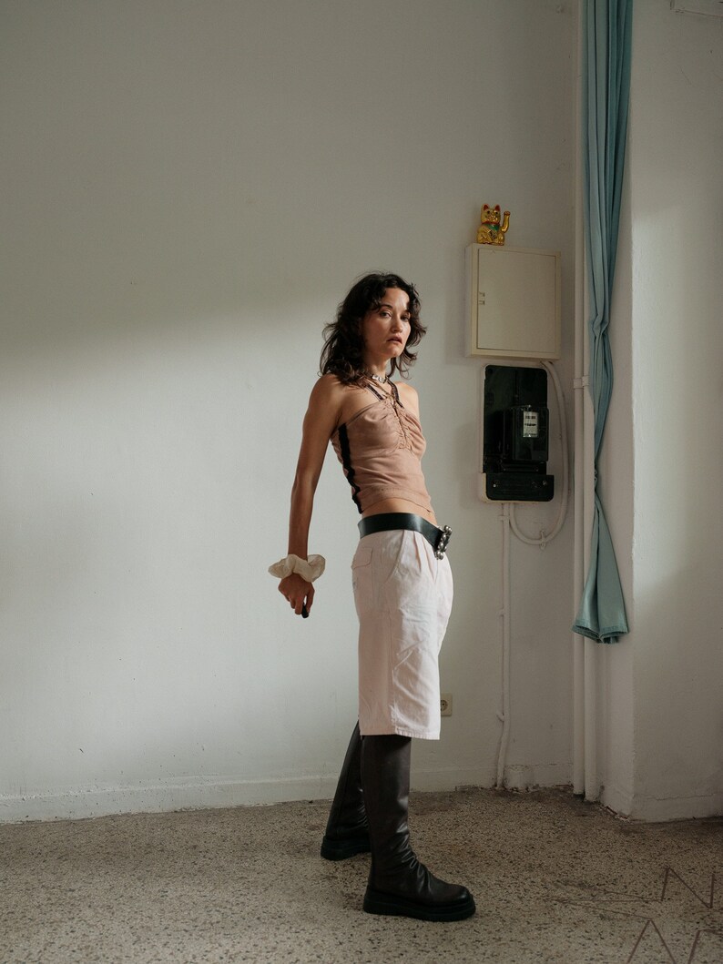 May include: A woman wearing a brown halter top, a black belt, white shorts, and black knee-high boots. She is standing in a room with white walls and a blue curtain.