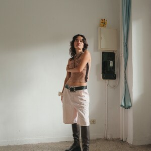May include: A woman wearing a brown tank top with a black lace trim, a black belt with a silver buckle, and light pink shorts. She is wearing black knee-high boots and has her arms crossed.