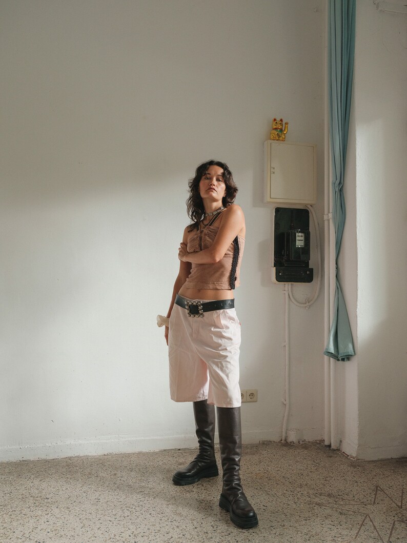 May include: A woman wearing a brown tank top with a black lace trim, a black belt with a silver buckle, and light pink shorts. She is wearing black knee-high boots and has her arms crossed.