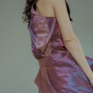 Op de afbeelding: Een vrouw draagt een mouwloze, iriserende jurk in paars en roze met een halternek. De jurk heeft een aansluitend bovenstuk en een zwierige rok.