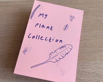 May include: A pink notebook with the words "My Plant Collection" written in blue ink. The cover features a drawn feather and leaf, along with small dotted designs. The notebook is on a light wood surface.