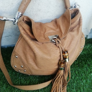 May include: A tan leather handbag with a long strap and a bamboo tassel. The bag has a textured leather exterior and silver hardware.