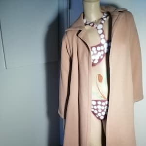 Puede incluir: Un abrigo largo de color marrón claro con cuello y mangas largas. El abrigo se exhibe en un maniquí, revelando un traje de baño estampado debajo. El maniquí está colocado frente a una pared azul claro.