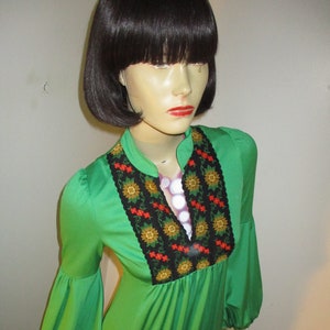 May include: A green maxi dress with long sleeves and a floral embroidered neckline. The dress has a gathered waist and a flowing skirt.