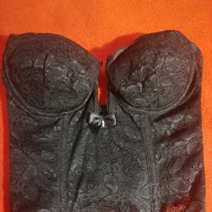 May include: Black lace corset with a sweetheart neckline and a small bow detail. The corset has a structured design with molded cups and a fitted waist, showcasing a floral pattern.