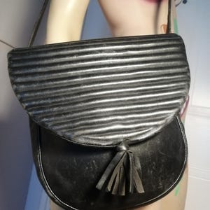 Puede incluir: Bolso de hombro de cuero negro con solapa plateada con costuras horizontales. El bolso tiene una parte inferior curvada y un detalle de borla. La correa es fina y negra. El bolso se exhibe en un maniquí.