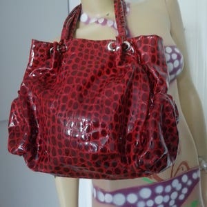 May include: A glossy red handbag featuring a textured crocodile-skin pattern. The bag has two top handles and silver-toned metal grommets at the top opening, which allow for a gathered, slouchy silhouette. The material has a shiny, reflective finish.