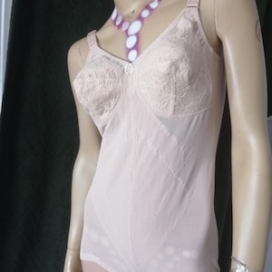 May include: A light beige bodysuit featuring lace-detailed cups with a small central floral accent, wide shoulder straps, and structured vertical seams. The garment is constructed from a smooth, opaque fabric with a fitted silhouette and a scalloped hem.
