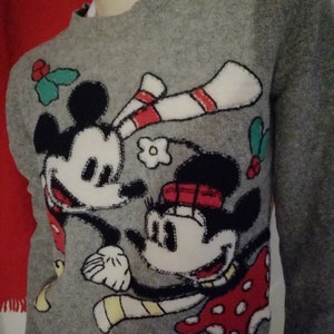 Peut inclure: Un pull gris avec Mickey et Minnie Mouse. Le motif comprend des contours noirs, des accents blancs, rouges et jaunes, et quelques feuilles de houx vertes. Le pull a un col rond et des manches longues.