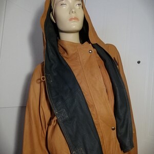 Peut inclure: Veste en cuir marron clair avec capuche et détails noirs. La veste a une fermeture à boutons et une taille ajustable par cordon. La capuche est doublée de tissu noir. La veste est présentée sur un mannequin.