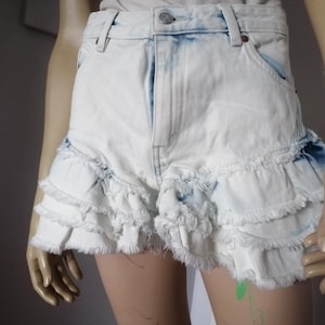 Può includere: Pantaloncini di jeans bianchi con orlo a balze. I pantaloncini hanno un lavaggio blu chiaro e bordi sfilacciati. Hanno un classico design a cinque tasche e una chiusura con bottoni.