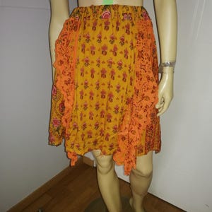 Peut inclure: Jupe jaune moutarde avec un motif floral et une bordure en dentelle orange. La jupe a un design à volants et est présentée sur un mannequin. La jupe est de style genou.