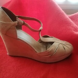 Puede incluir: Sandalia de cuña color beige con diseño de tira en T. La sandalia presenta una suela de plataforma y una puntera cerrada con pliegues decorativos. El zapato está sobre un fondo rojo.