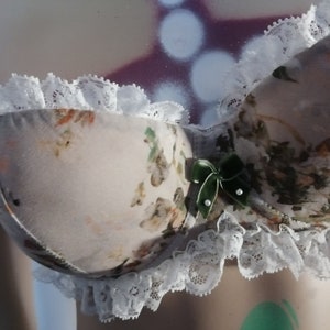 Könnte beinhalten: Ein BH mit Blumenmuster, weißer Spitzenborte und einer grünen Schleife. Der BH hat ein zartes, feminines Design.