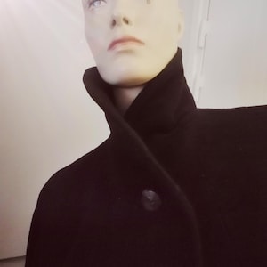 Puede incluir: Un abrigo negro con cuello alto y cierre de un solo botón. El abrigo se exhibe en un maniquí con tez pálida y maquillaje sutil. La tela del abrigo parece ser de un color oscuro y liso.