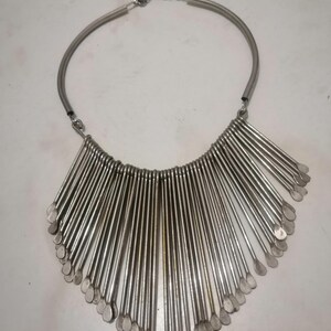 May include: A silver-toned necklace featuring a rigid, curved collar and a fan-shaped pendant. The pendant is composed of numerous vertical, elongated metal rods, each ending in a flattened, teardrop-shaped tip. The necklace is designed to be worn around the neck.
