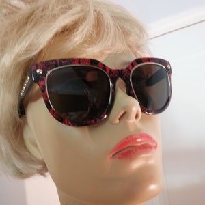 May include: Red and black patterned sunglasses with a silver frame. The sunglasses are on a mannequin head with blonde hair.