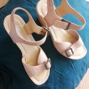 Peut inclure: Sandales en cuir marron clair avec des lanières réglables et des boucles métalliques. Les sandales ont un talon compensé. Le nom de la marque "Clarks ORIGINALS" est visible sur le côté. Elles sont posées sur un tissu bleu canard.