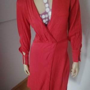Könnte beinhalten: Ein rotes Wickelkleid mit langen Ärmeln und einem V-Ausschnitt. Das Kleid hat einen strukturierten Stoff und einen leicht ausgestellten Rock.